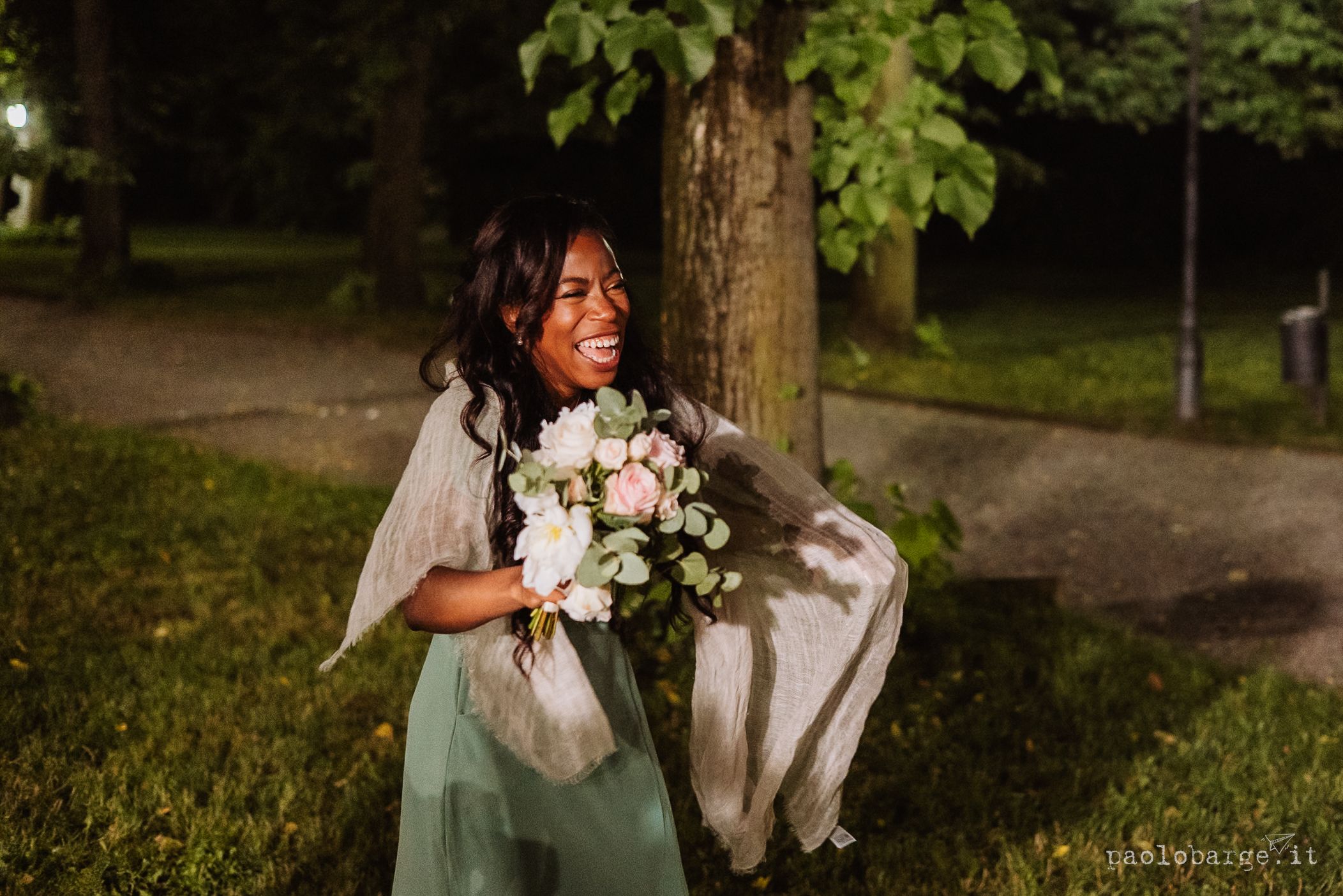A smiling person in a mint green dress holds a white bouquet of flowers while standing near a tree in a park setting.