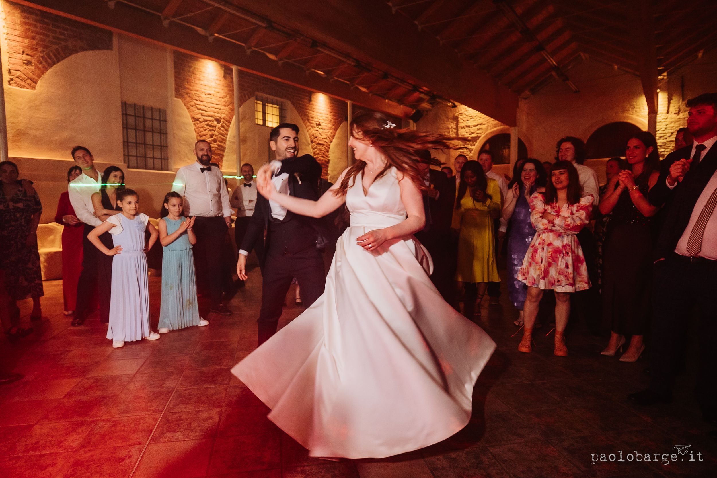 Wedding reception guests dance and celebrate on a rustic wooden floor under warm ambient lighting.
