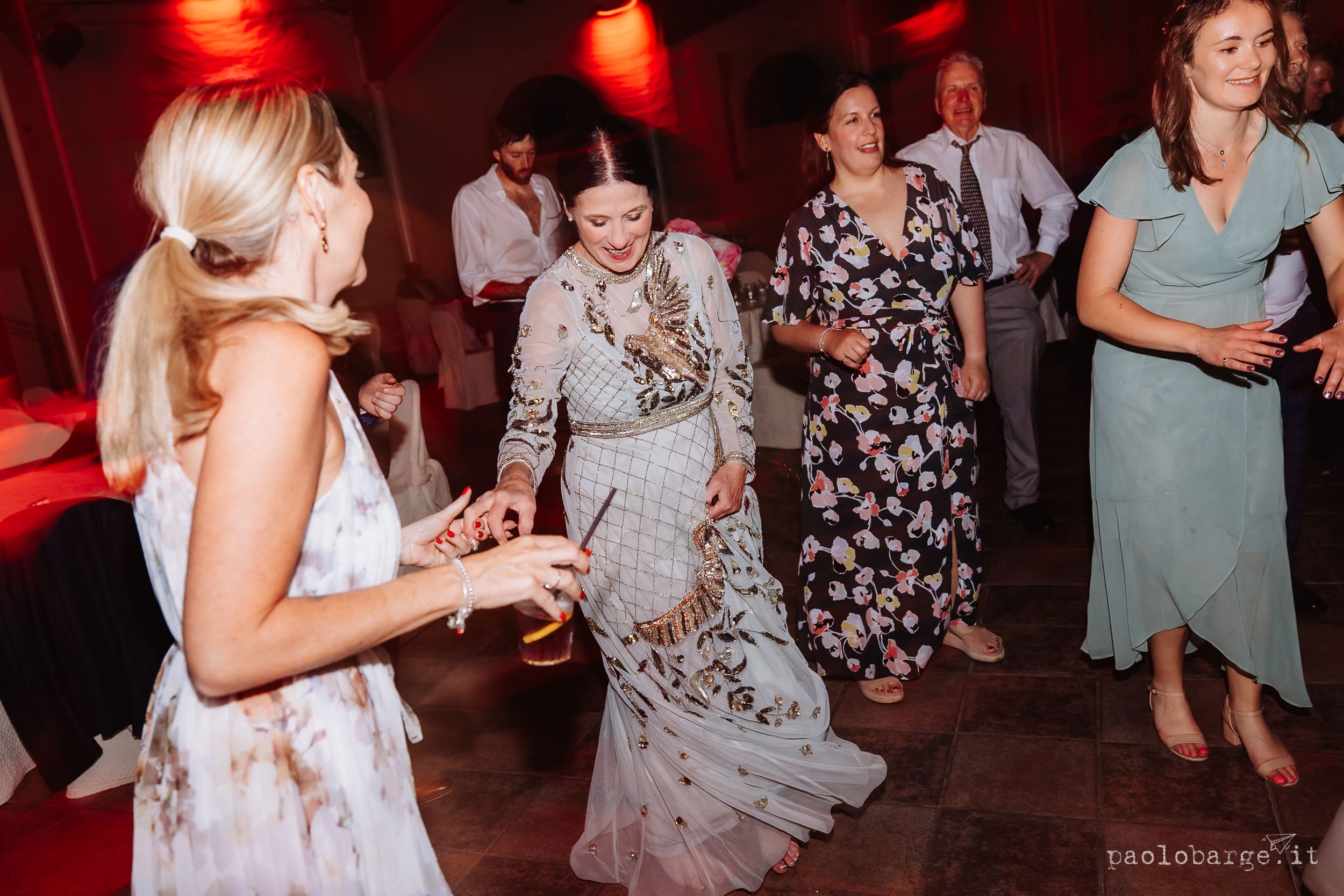 People dancing and celebrating at a wedding reception with red lighting and bright formal dresses on dance floor.