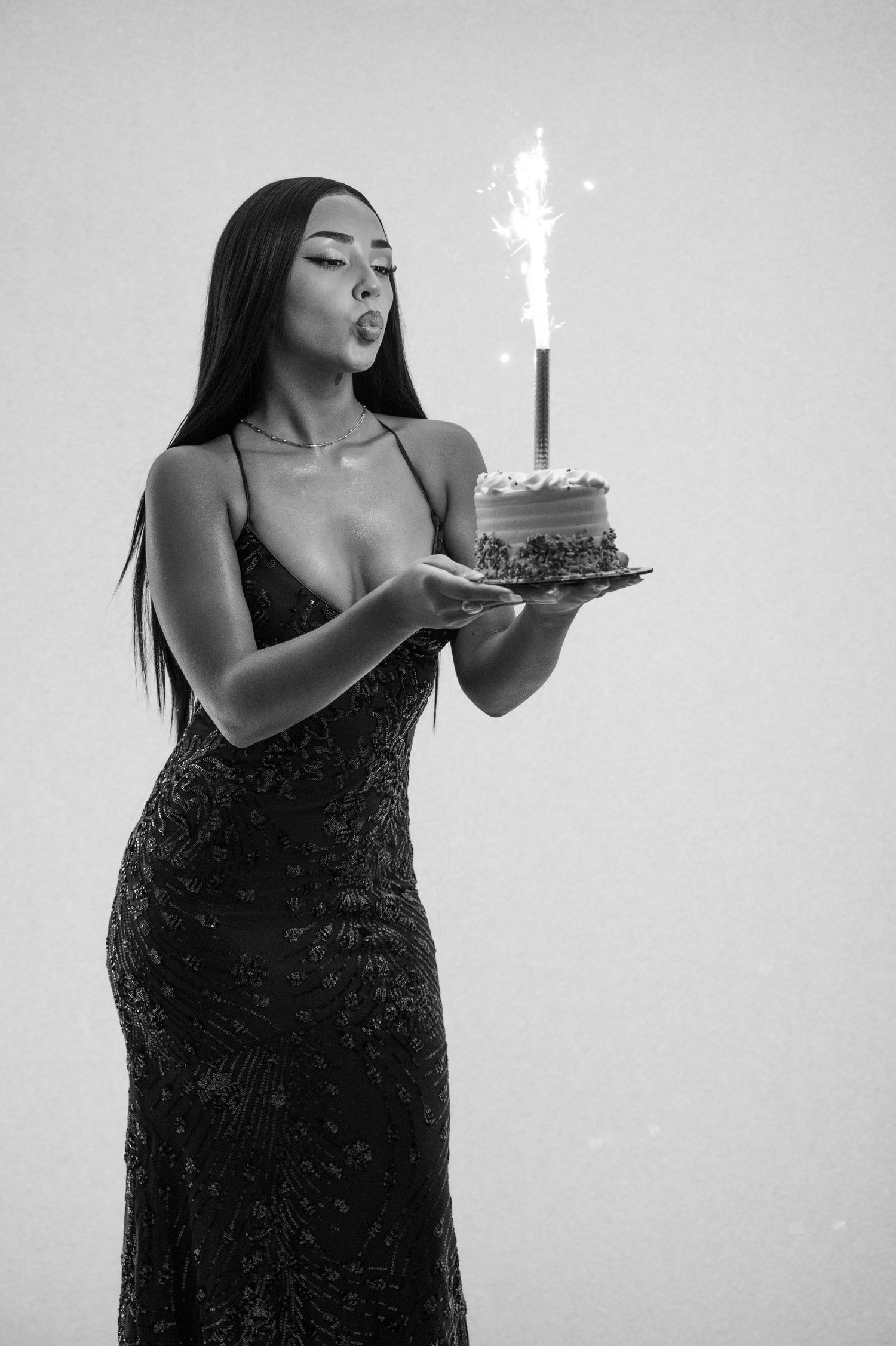 A person in a black sequined evening dress poses with a birthday cake lit with sparklers in a studio setting.