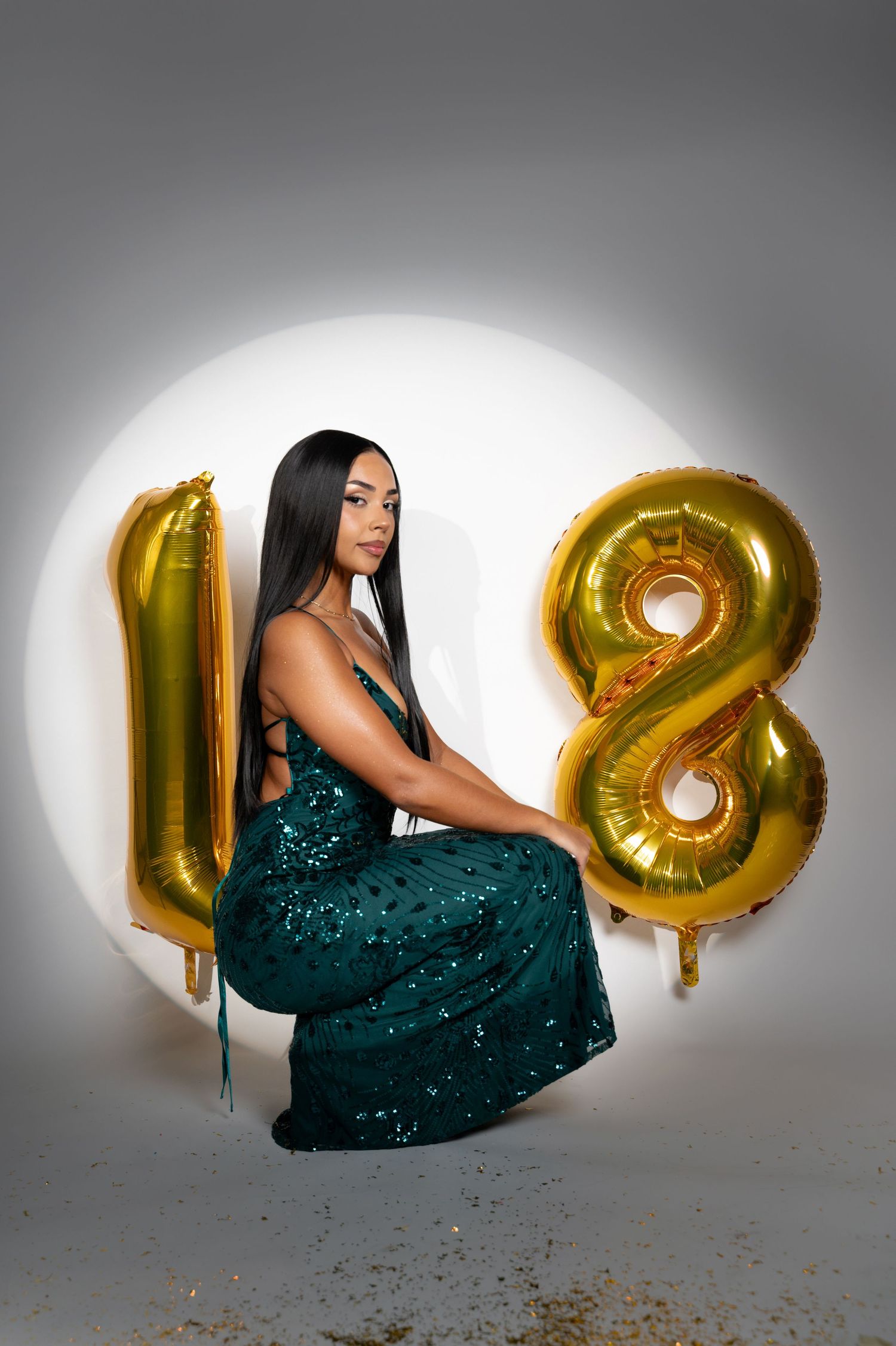 A figure in a sparkling emerald green gown poses next to large gold number balloons showing 18 against a white backdrop.