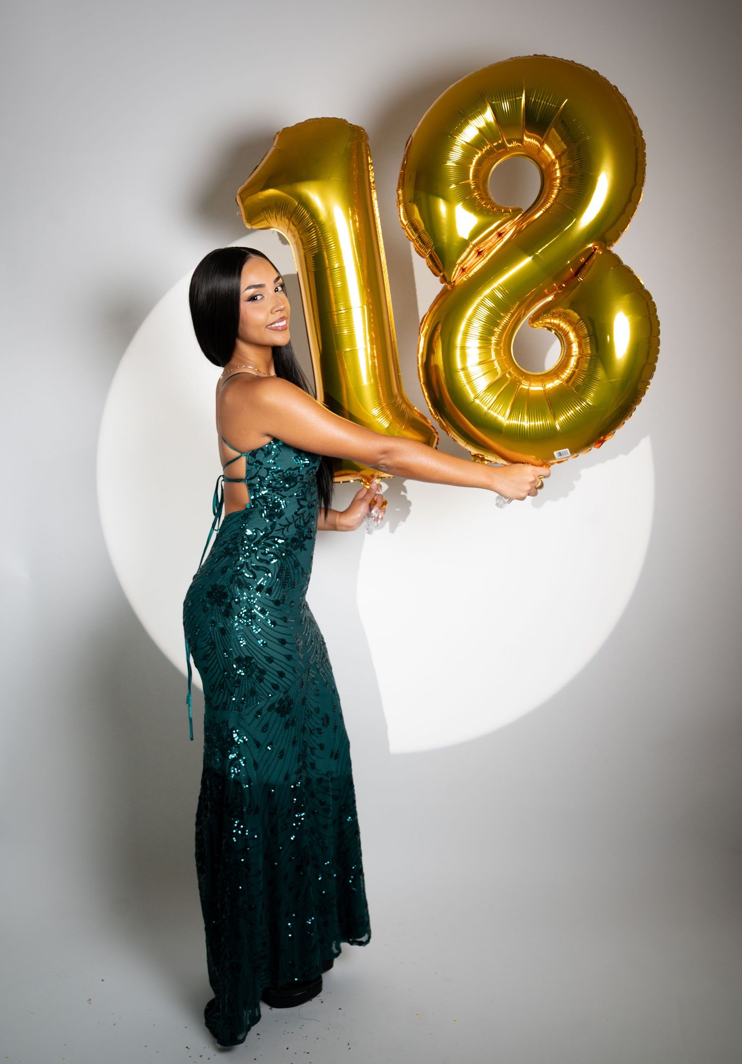 Figure in a sparkling emerald green formal dress poses with gold number 18 balloons against a white backdrop.