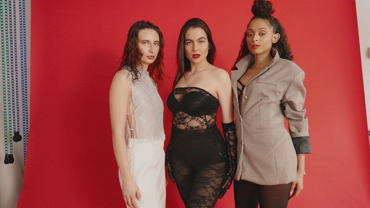 Three women pose together against a red backdrop wearing contrasting formal outfits in white, black lace, and tan colors.
