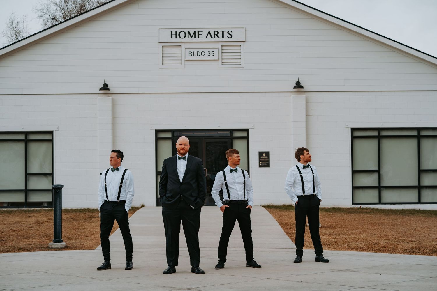 Group in dark suits and suspenders stand in line against white building exterior for formal wedding photo.