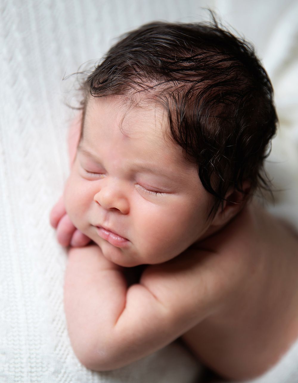 A sleeping newborn baby with dark hair rests peacefully against a white textured background. During a newborn session in London
