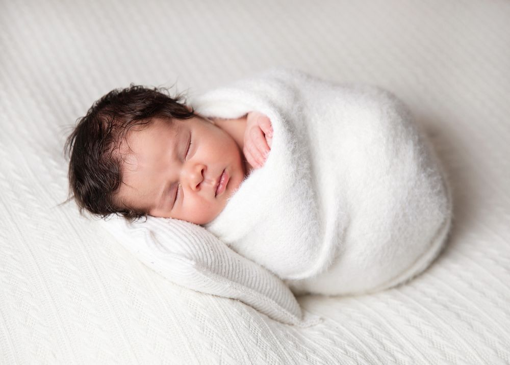 Peaceful sleeping newborn baby swaddled in soft white blanket rests on light textured surface.