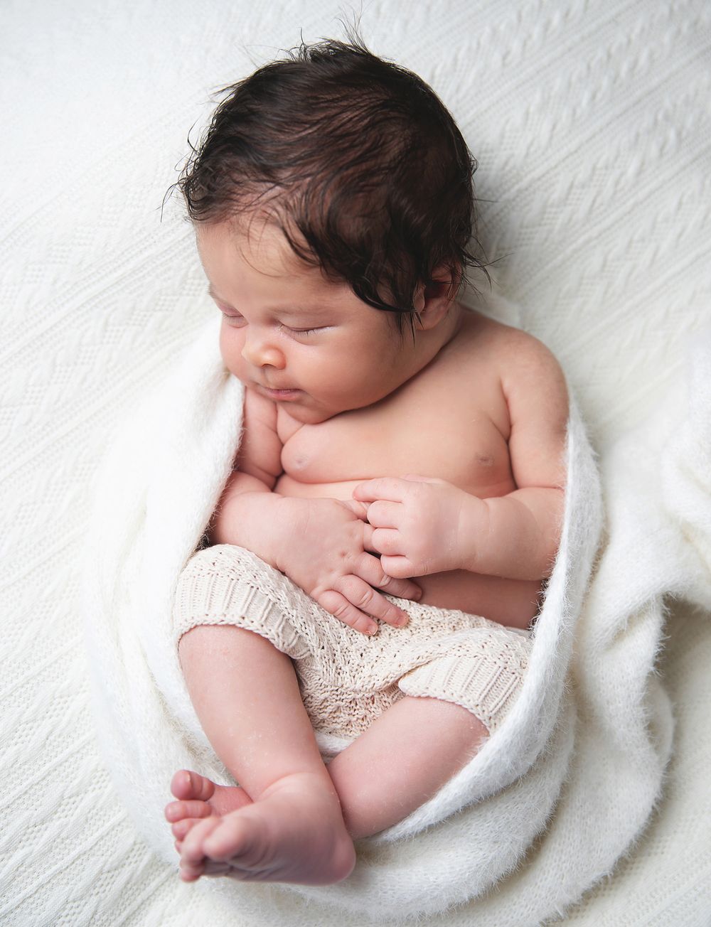 A newborn baby sleeps peacefully wrapped in white fabric with a knit accessory during a professional photoshoot.