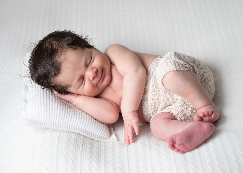 Sleeping newborn baby wearing white lace diaper cover rests on soft white pillow with peaceful expression. During a newborn session.