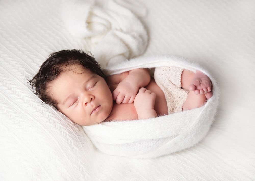 Sleeping newborn baby wrapped in soft white blanket on white textured surface. During a newborn family session in London