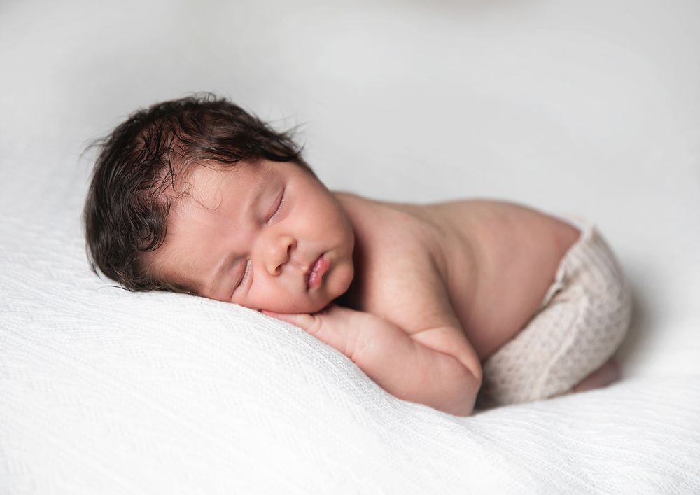 A peaceful newborn baby sleeping soundly on white fabric wearing cream-colored swaddle wrap. During a newborn family session in London.