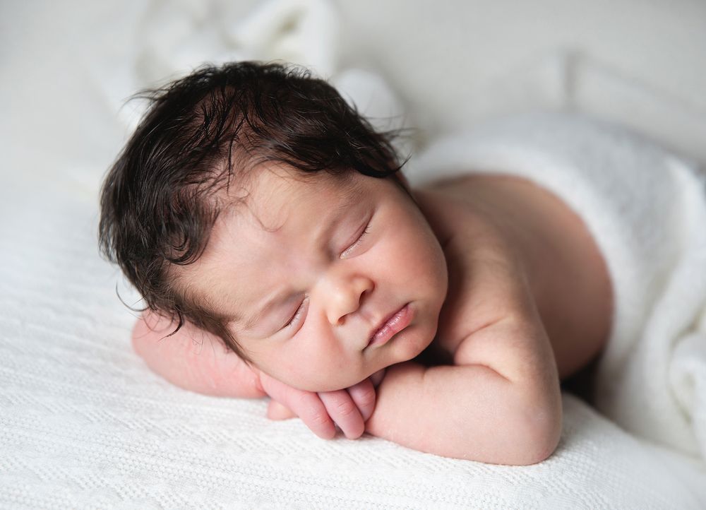 A peaceful sleeping newborn baby lies on soft white fabric with hands folded under their cheek. During a newborn family session in London