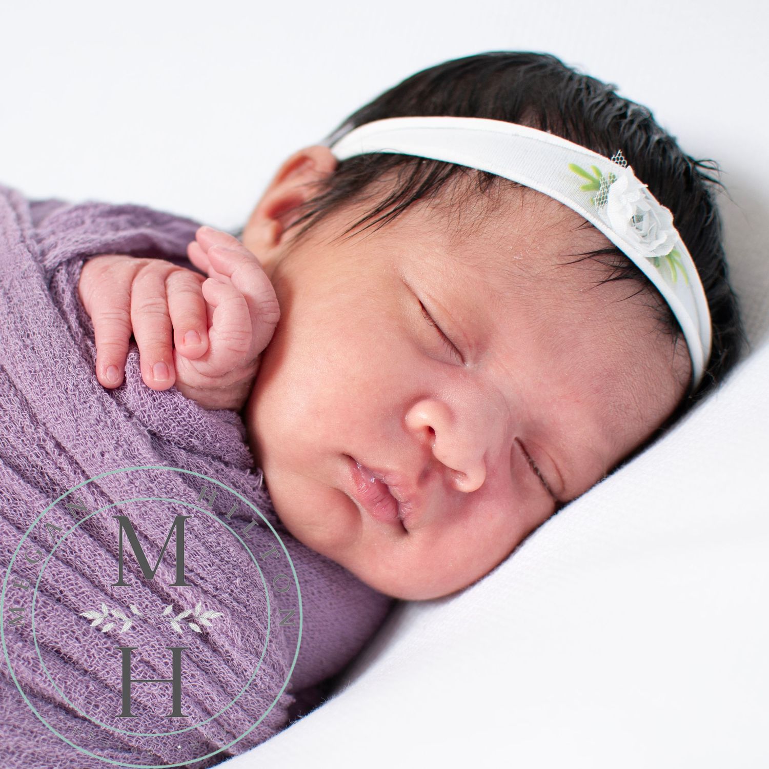Close-up profile view of a sleeping baby with black hair wearing a lavender swaddle and white headband on a light background.
