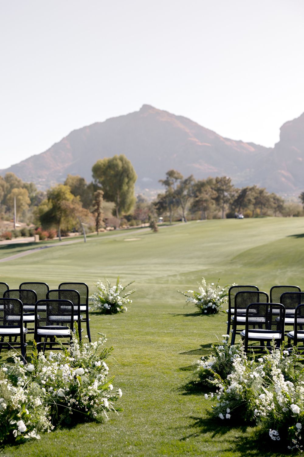 Outdoor wedding ceremony setup with white chairs and floral arrangements on a lush golf course with mountain views.