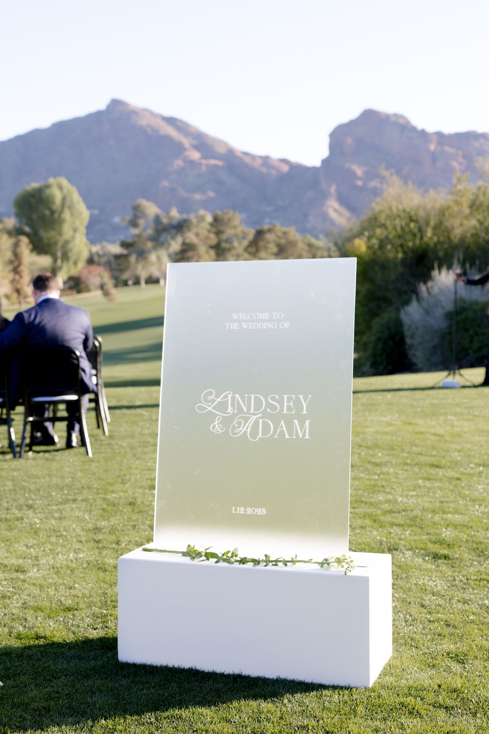 An elegant wedding welcome sign with Lindsey & Adam text displayed on a lawn with mountains in the background.