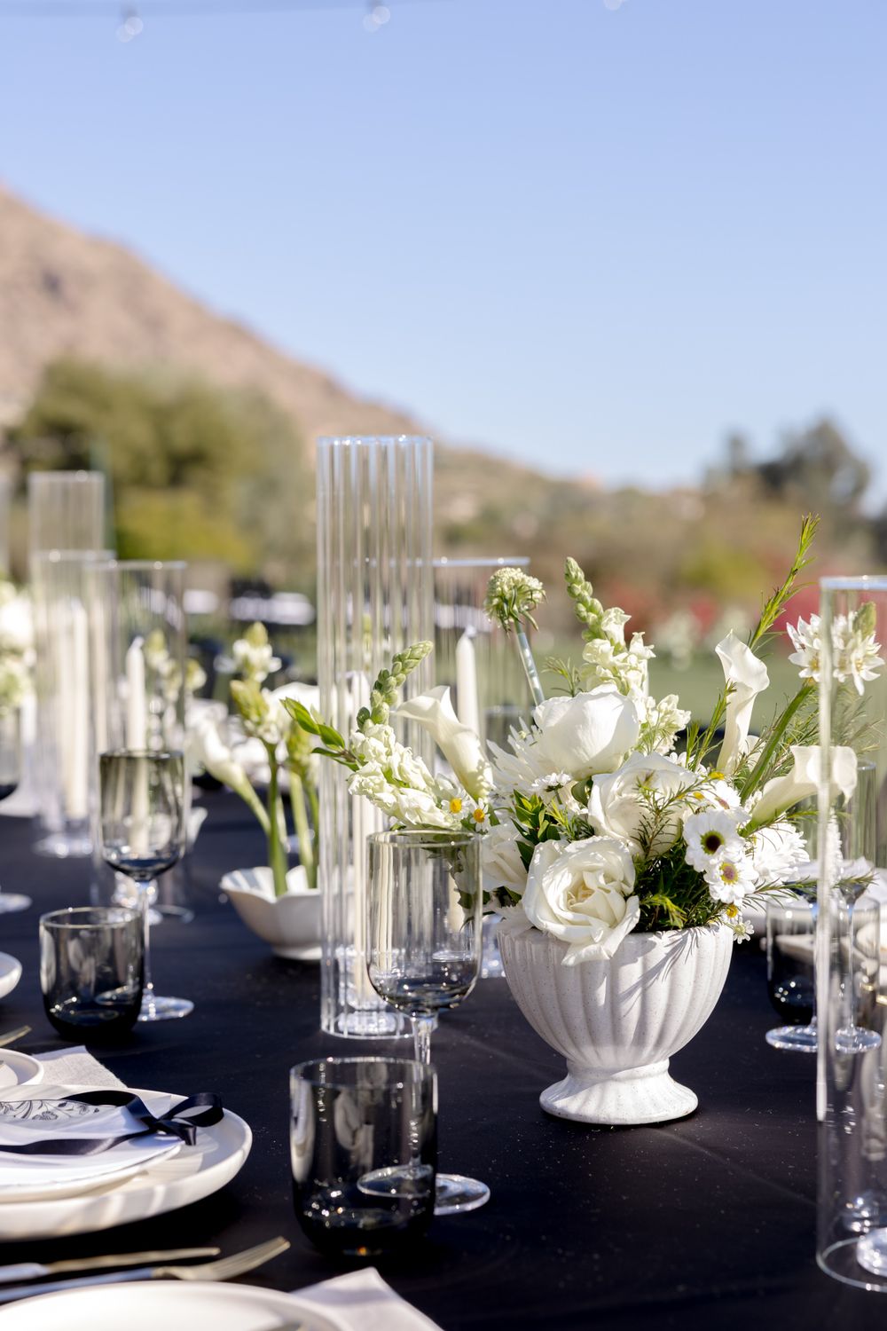 Elegant table setting with white floral centerpieces, tall glass vases, and black tablecloth against desert mountain backdrop.