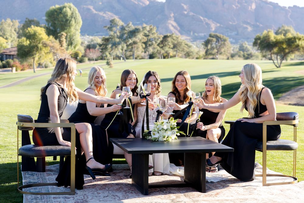 A group in black dresses gather outdoors for drinks and conversation at a table with mountain scenery in the background.
