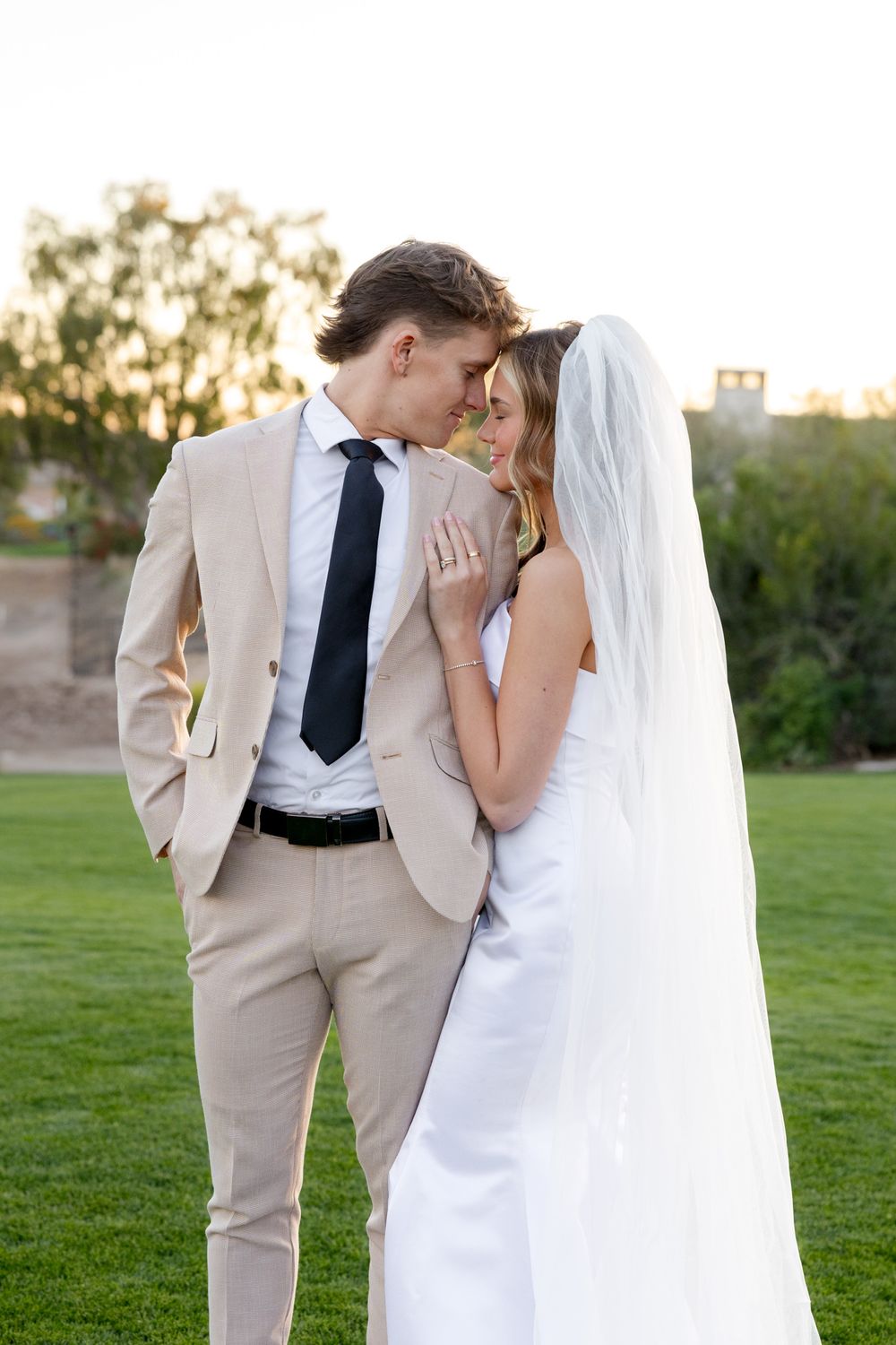 A romantic wedding portrait of a couple in a tan suit and white dress embracing on a grassy lawn at sunset.