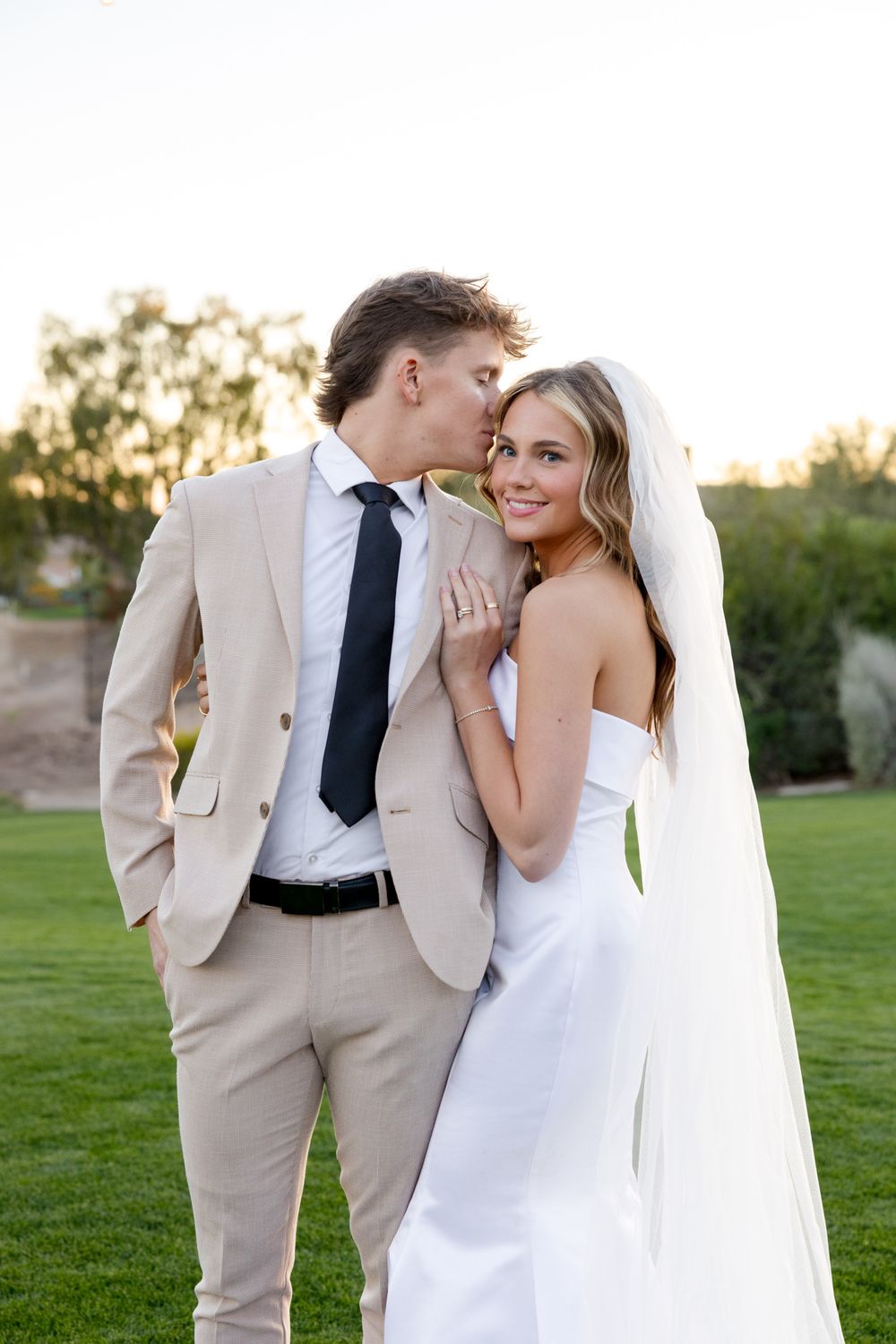 A romantic wedding photo of a couple in formal attire embracing on a lush lawn at sunset.