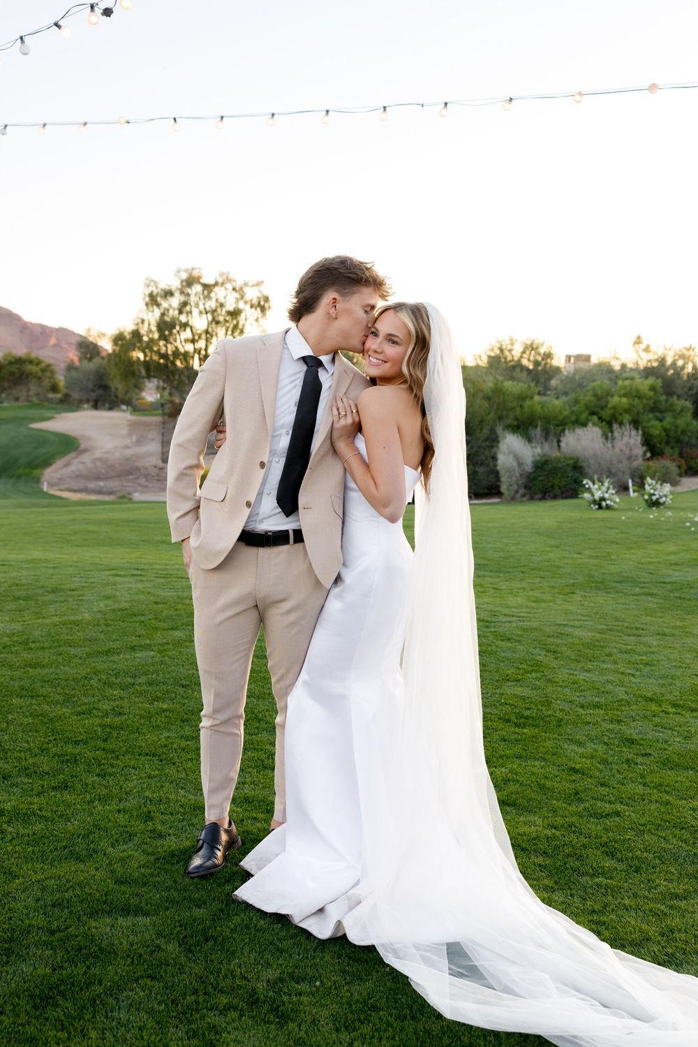 Wedding couple sharing intimate moments on a lawn with string lights and mountain backdrop at dusk.