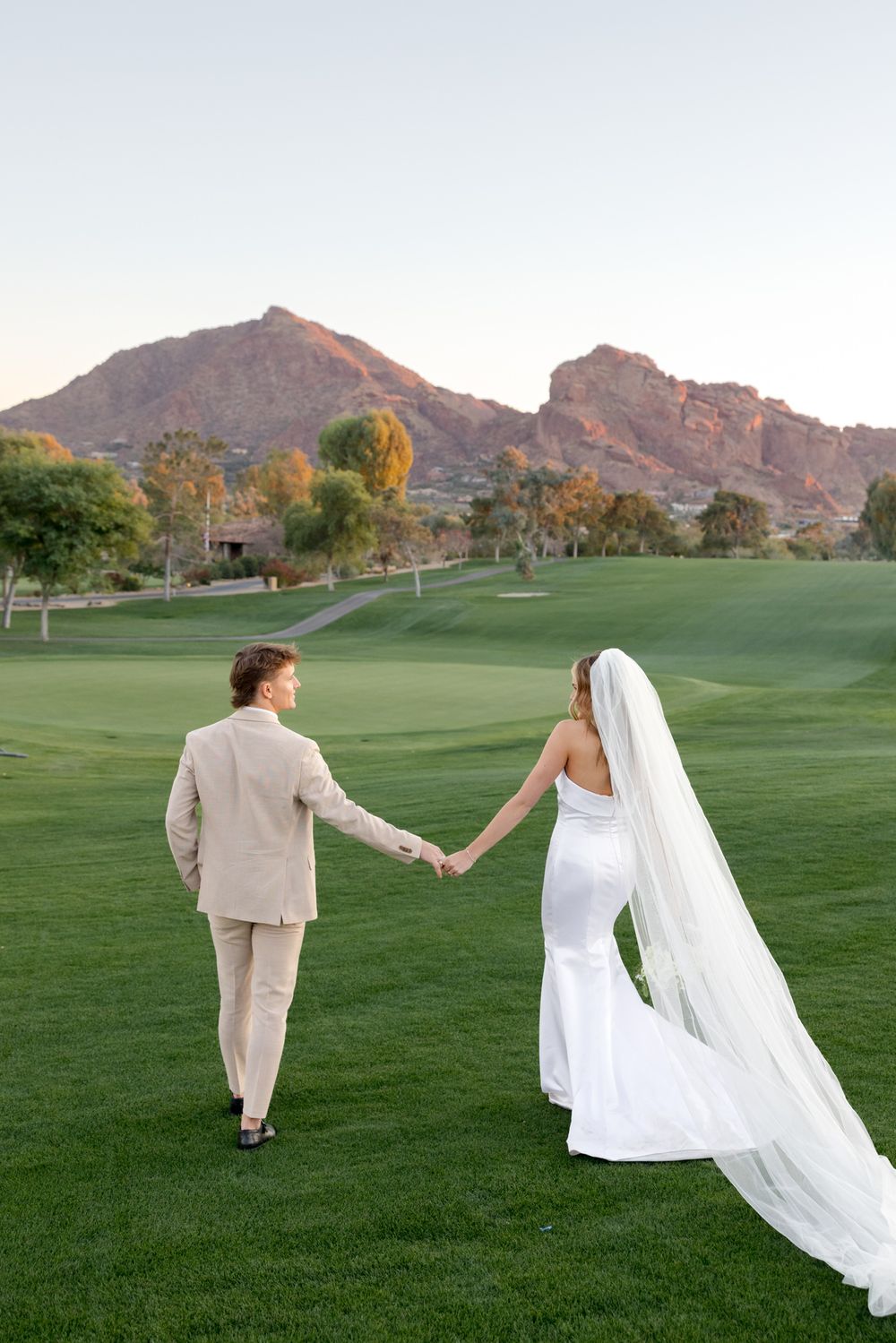 Couple walking hand in hand on golf course with dramatic mountain backdrop and flowing wedding veil.