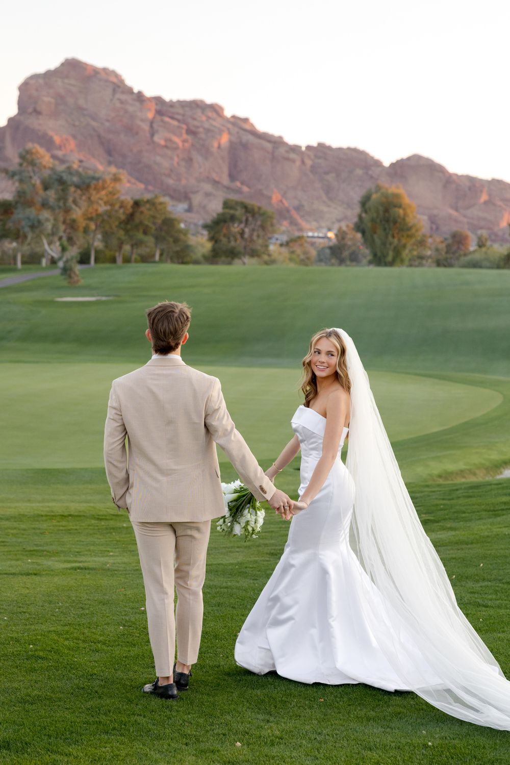 A couple poses for wedding photos on a golf course with dramatic mountain scenery behind them.