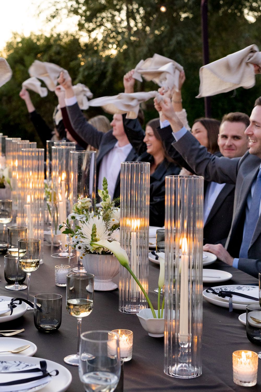 Wedding guests wave napkins in celebration at an elegant outdoor reception table with glowing candles.