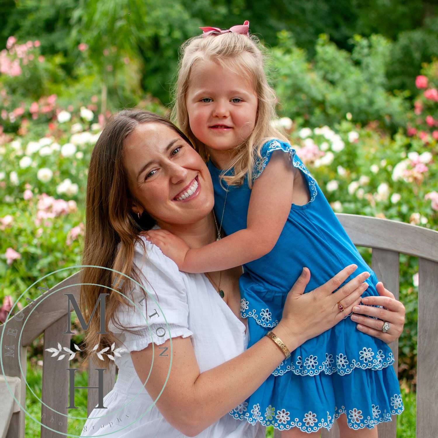 Woman and child in coordinating blue and white outfits embrace while sitting on wooden bench in garden setting.