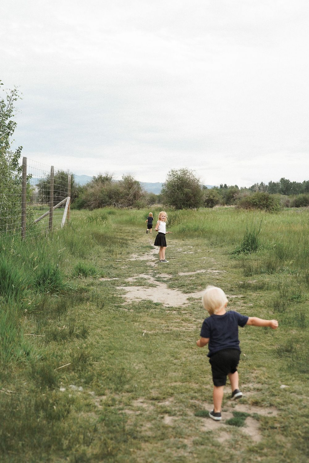 Two children in dark shorts running down a dirt path through a grassy meadow on a cloudy day.
