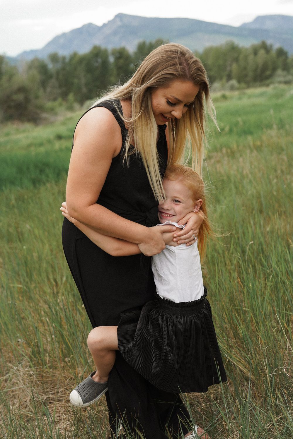 A tender moment shared between adult and child holding each other in a grassy meadow with mountains in background.