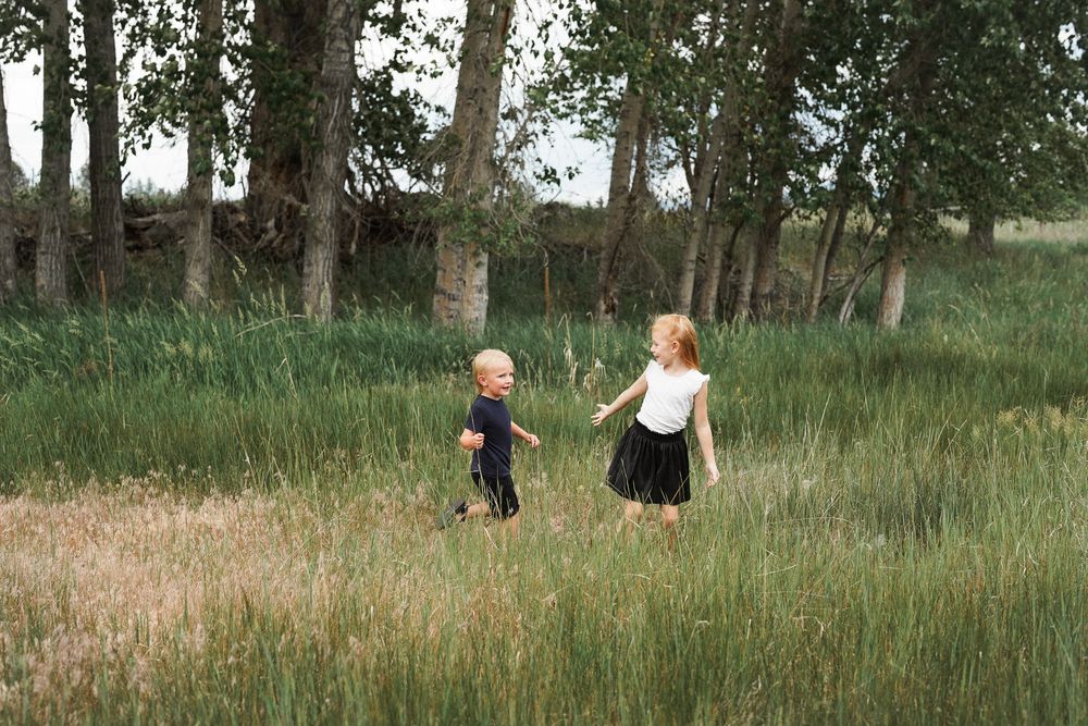 Two people walk through tall grass and wildflowers in a summer meadow while holding hands and walking away from the camera.