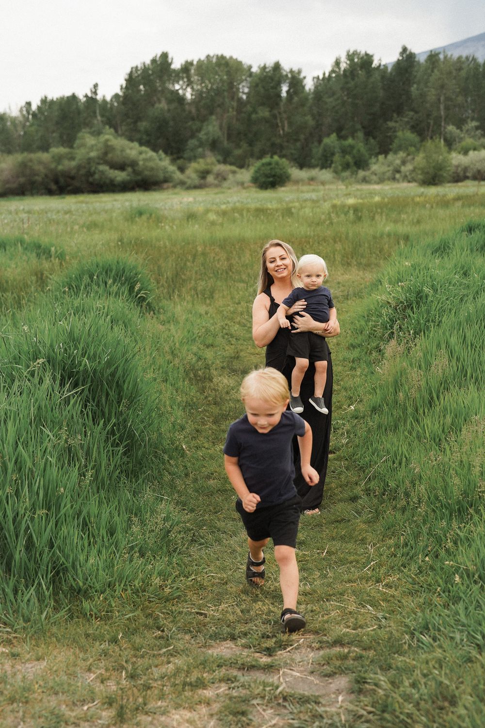 People walking along a dirt path through tall green grass with trees and mountains in the background.