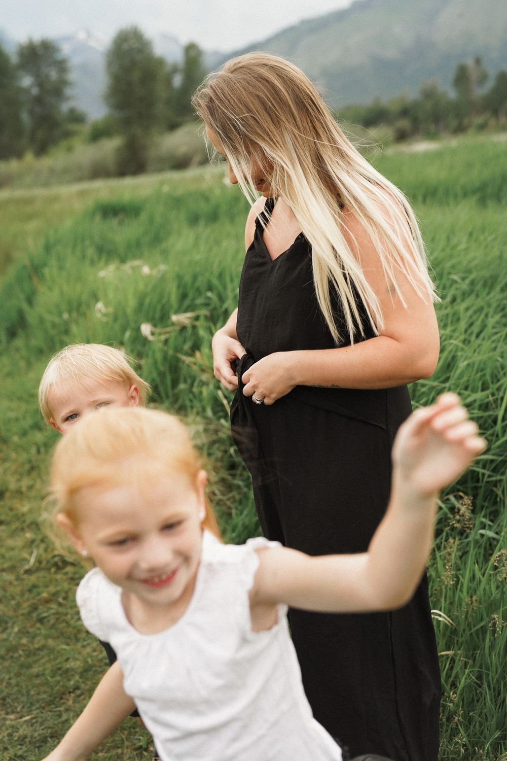 A pregnant woman in a black dress stands in a grassy field while a young child dances joyfully beside her.
