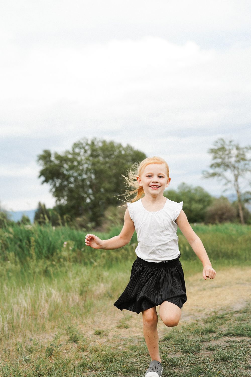 A child in a black skirt and white top runs joyfully through a lush green field with mountains in background.