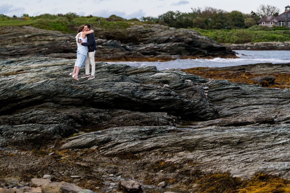 Seaside engagement photography on dramatic rocky coastline with waves crashing below.