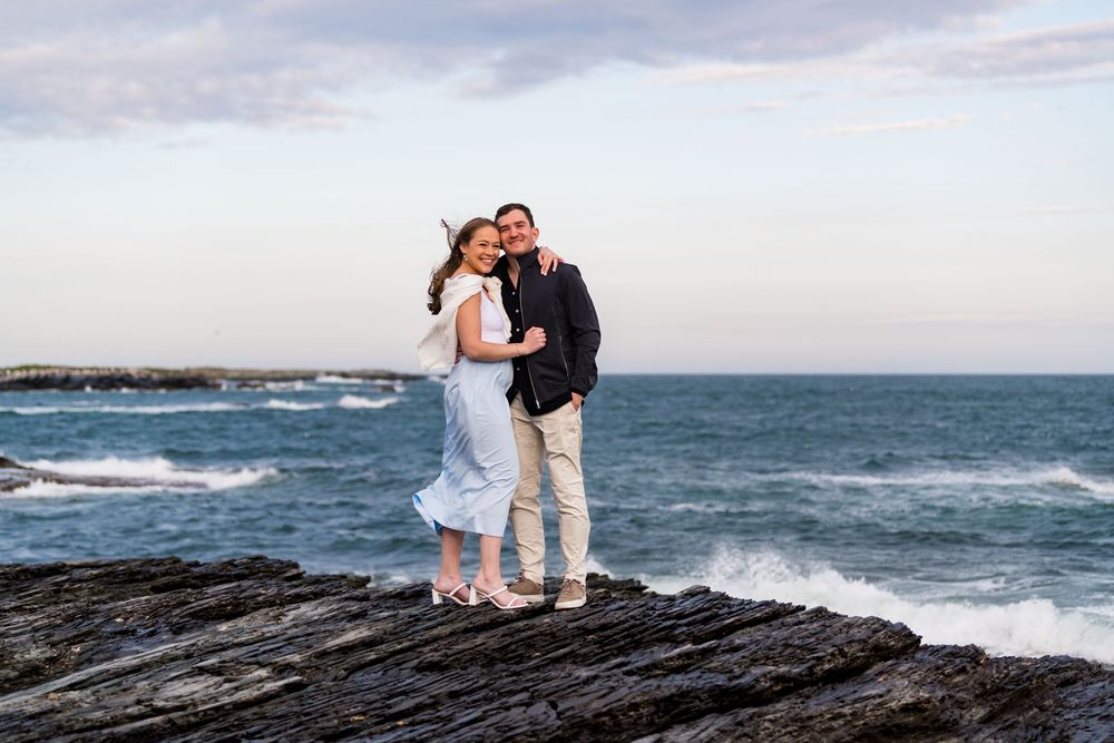 Couple embracing on rugged coastal rocks with ocean waves and cloudy sky in background.