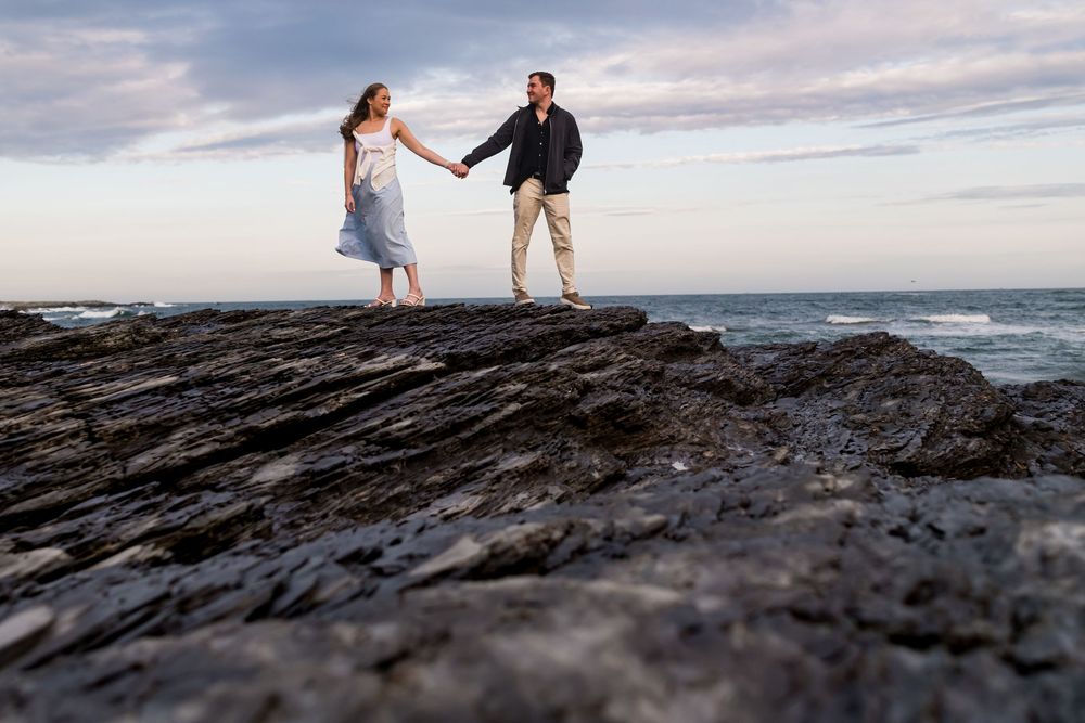A couple walks hand in hand along the dramatic rocky coastline against a moody cloudy sky in Hawaii.