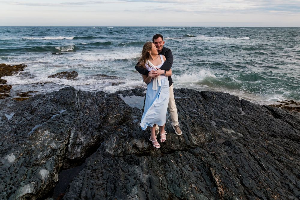 A romantic seaside embrace on dark rocky cliffs with crashing waves and ocean views in the background.