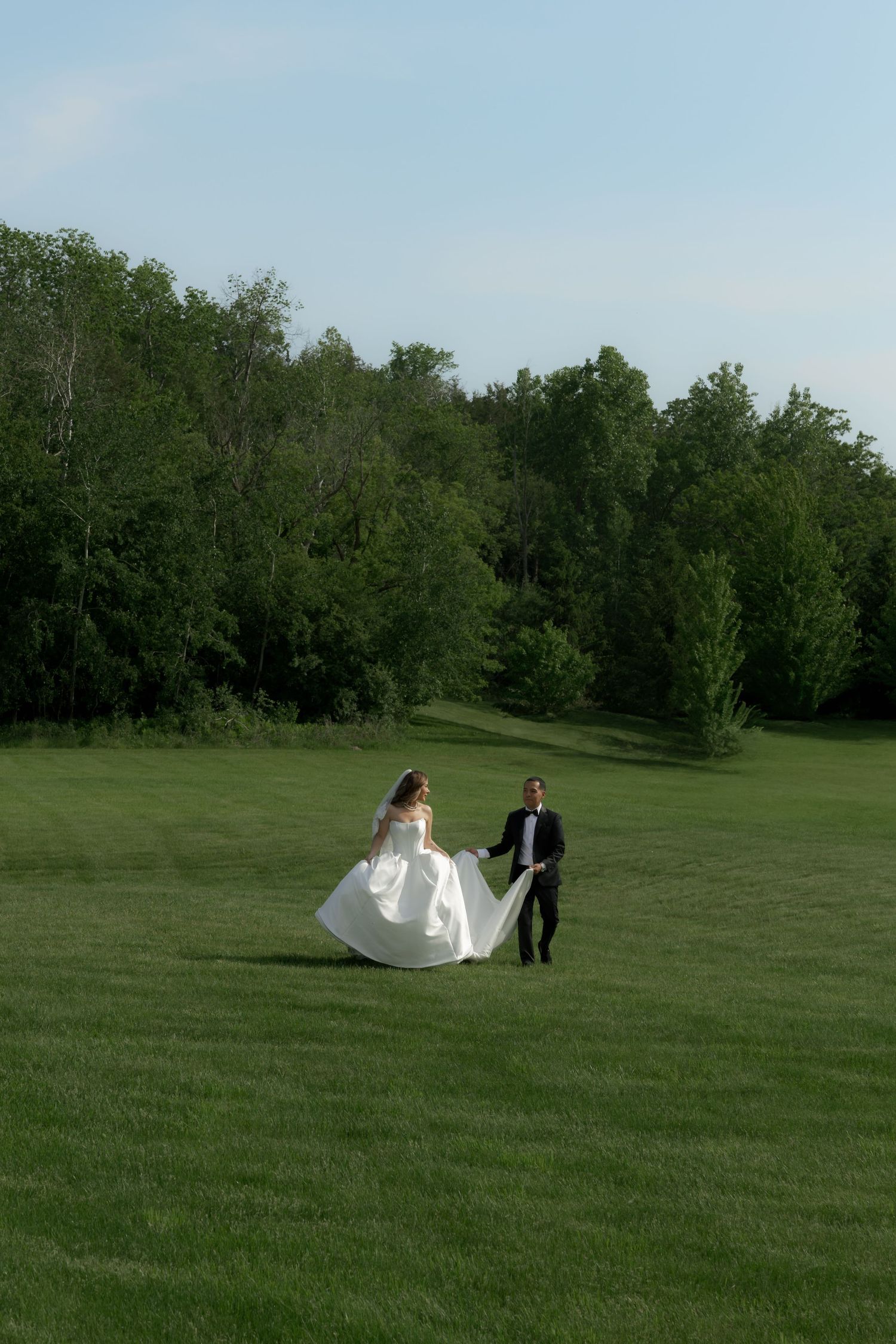 A couple in formalwear walks across a lush green lawn bordered by tall trees on a sunny day.