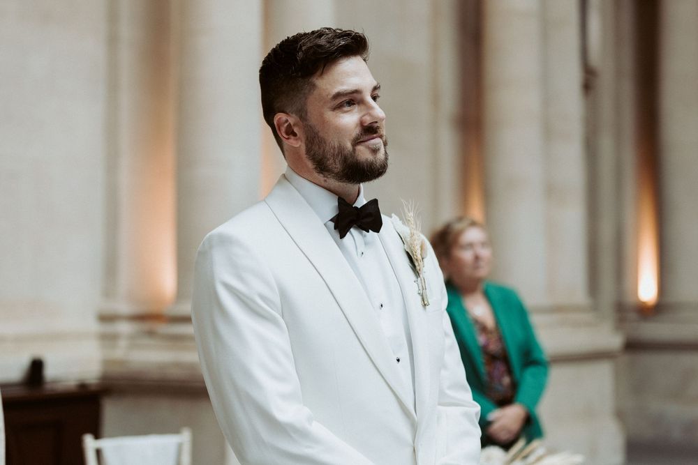 Groom in white tuxedo jacket and bow tie smiles while looking to the side during formal wedding event.
