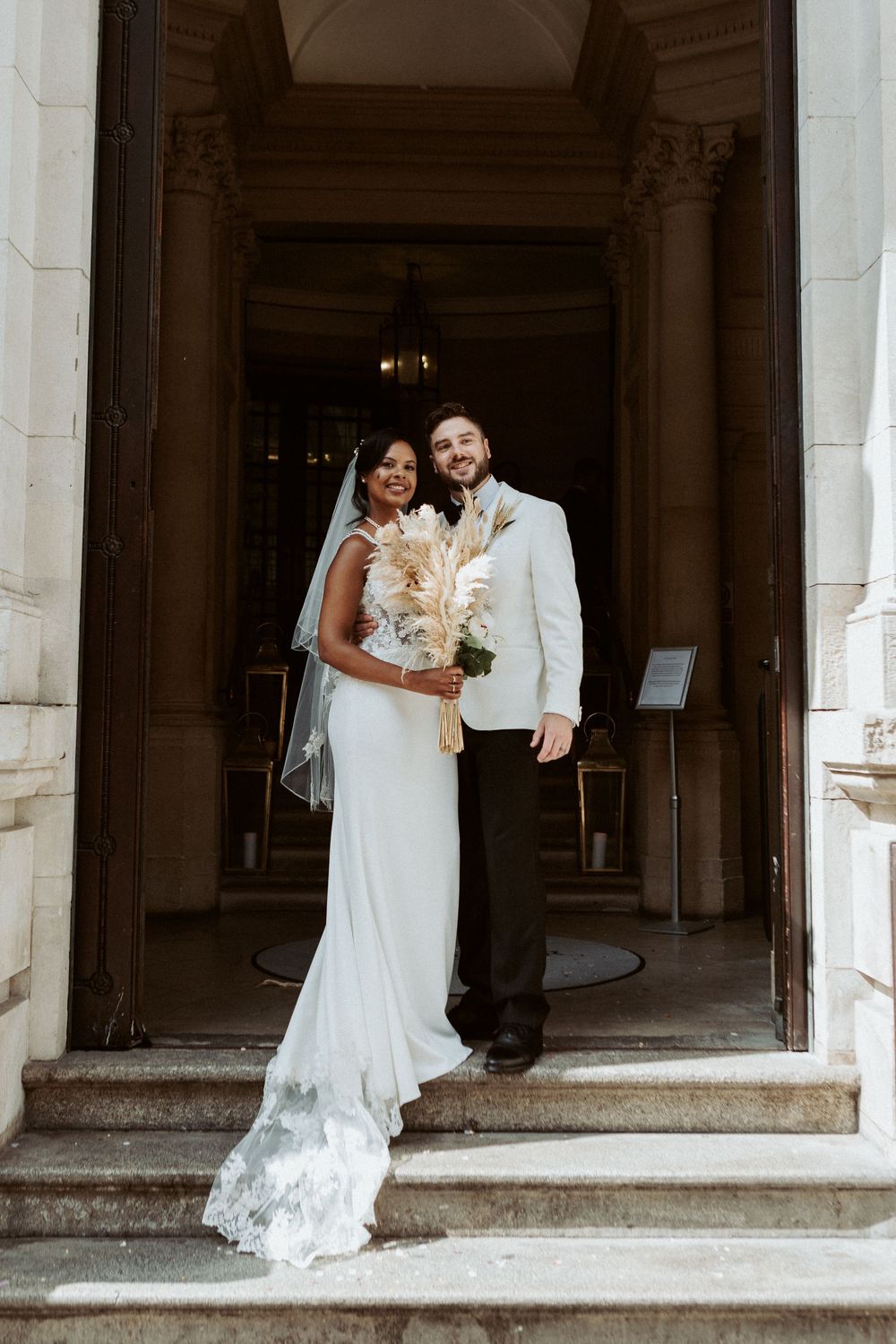 A couple stands on stone steps in wedding attire with the bride holding a pampas grass bouquet.