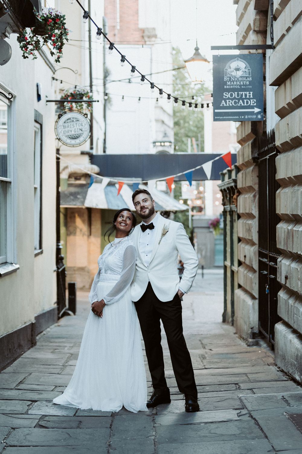A couple in wedding attire poses in a charming alleyway decorated with string lights and bunting in South Arcade.