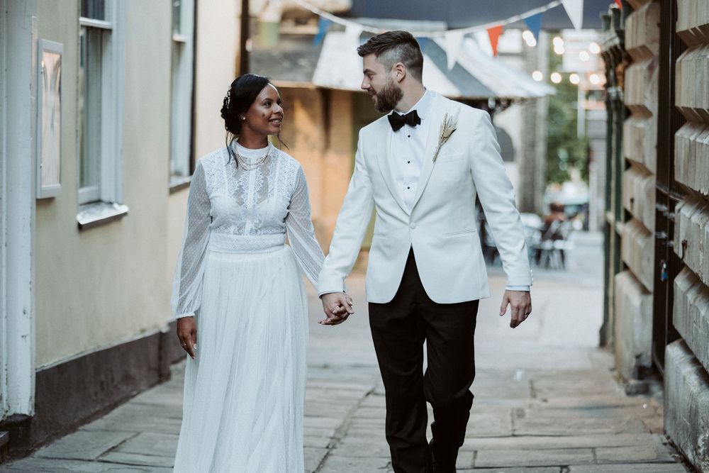 A couple in elegant wedding attire walks along a charming narrow cobblestone street in a European city.