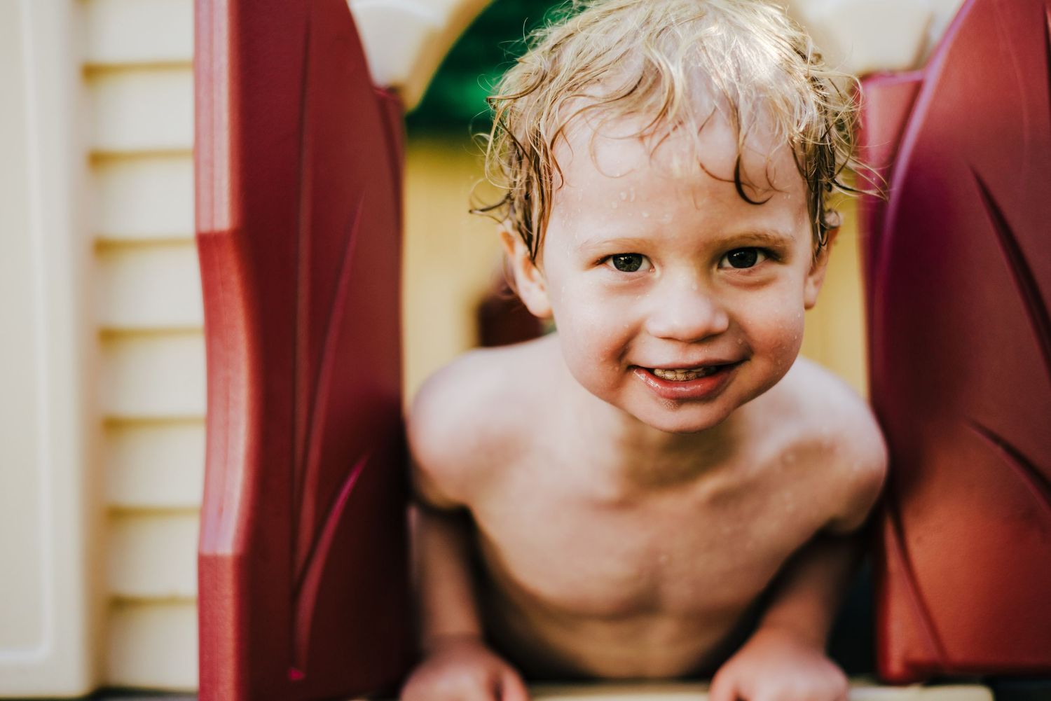 A child with water-slicked hair and bright expression peeks between playground equipment on a summer day.