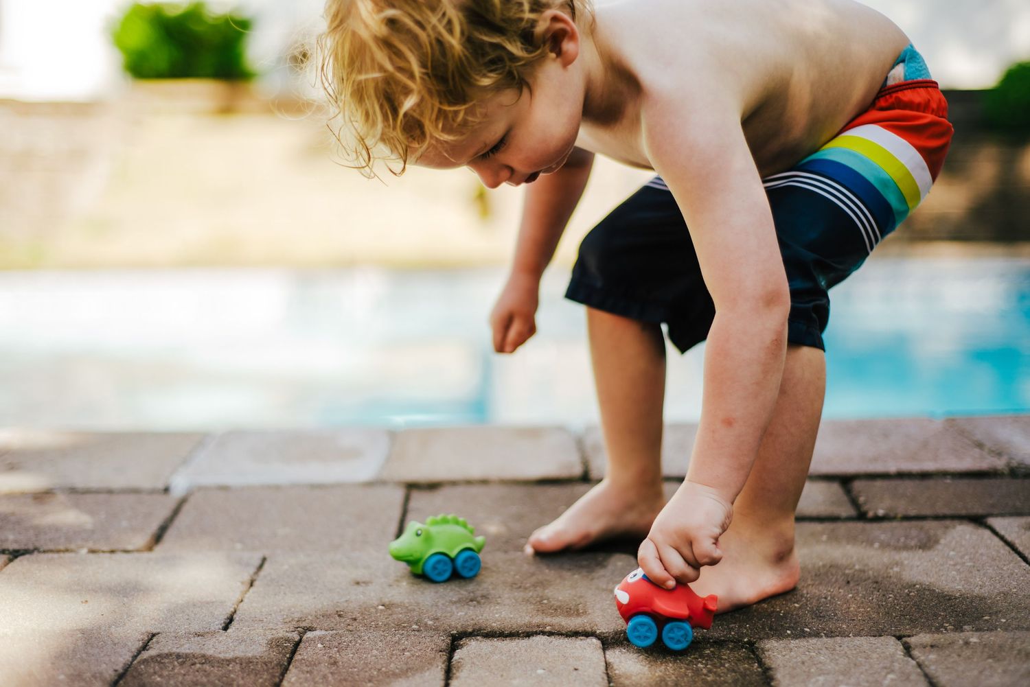 Child in colorful swim trunks bending down to play with toys by the edge of a swimming pool.