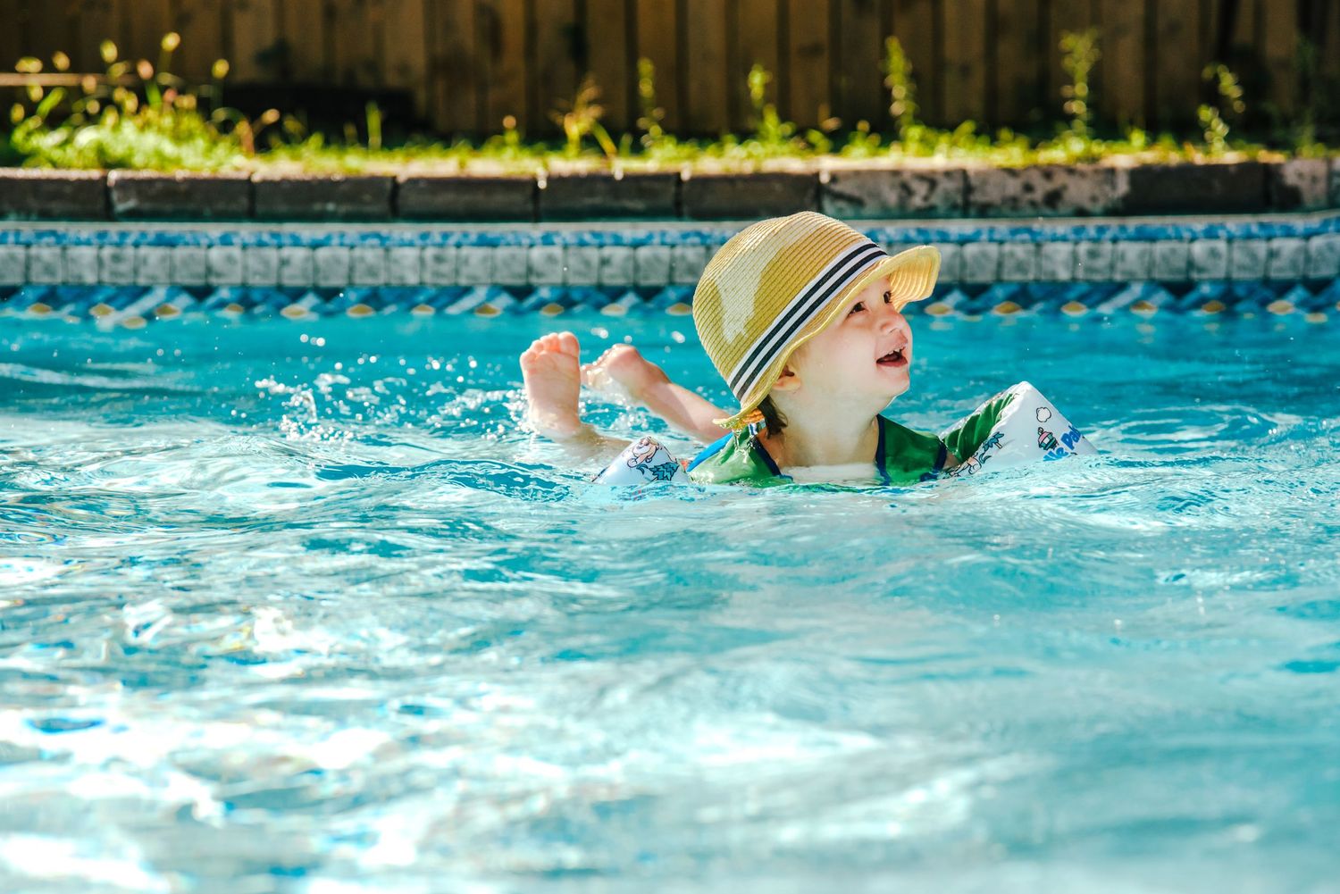 A swimmer wearing a sun hat splashes in a bright blue backyard swimming pool on a sunny day.