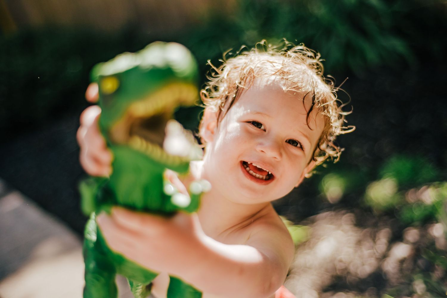 Young child playing with a green toy dinosaur outdoors on a sunny day.