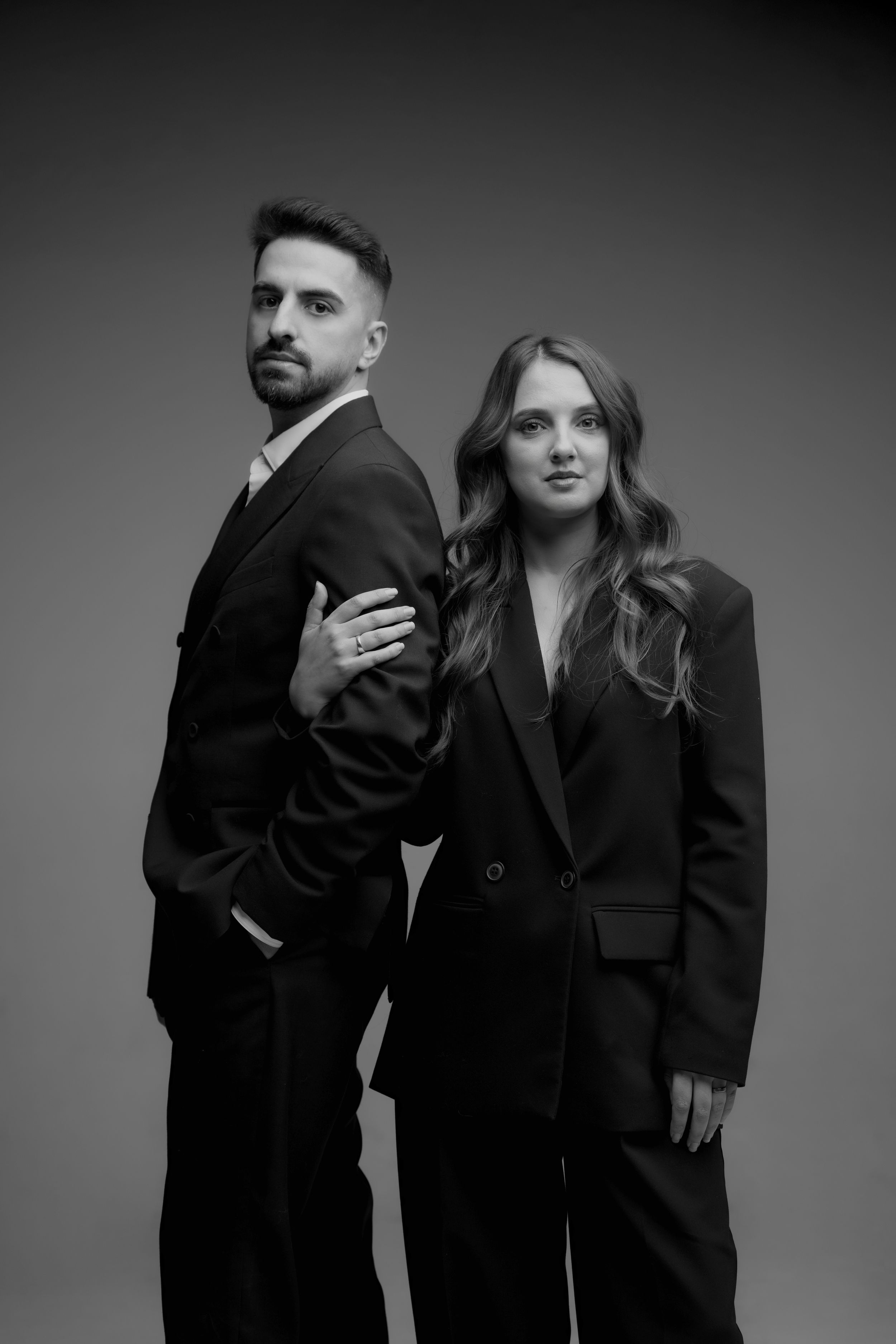 Stylish couple in matching black suits pose back-to-back in dramatic black and white studio portrait.