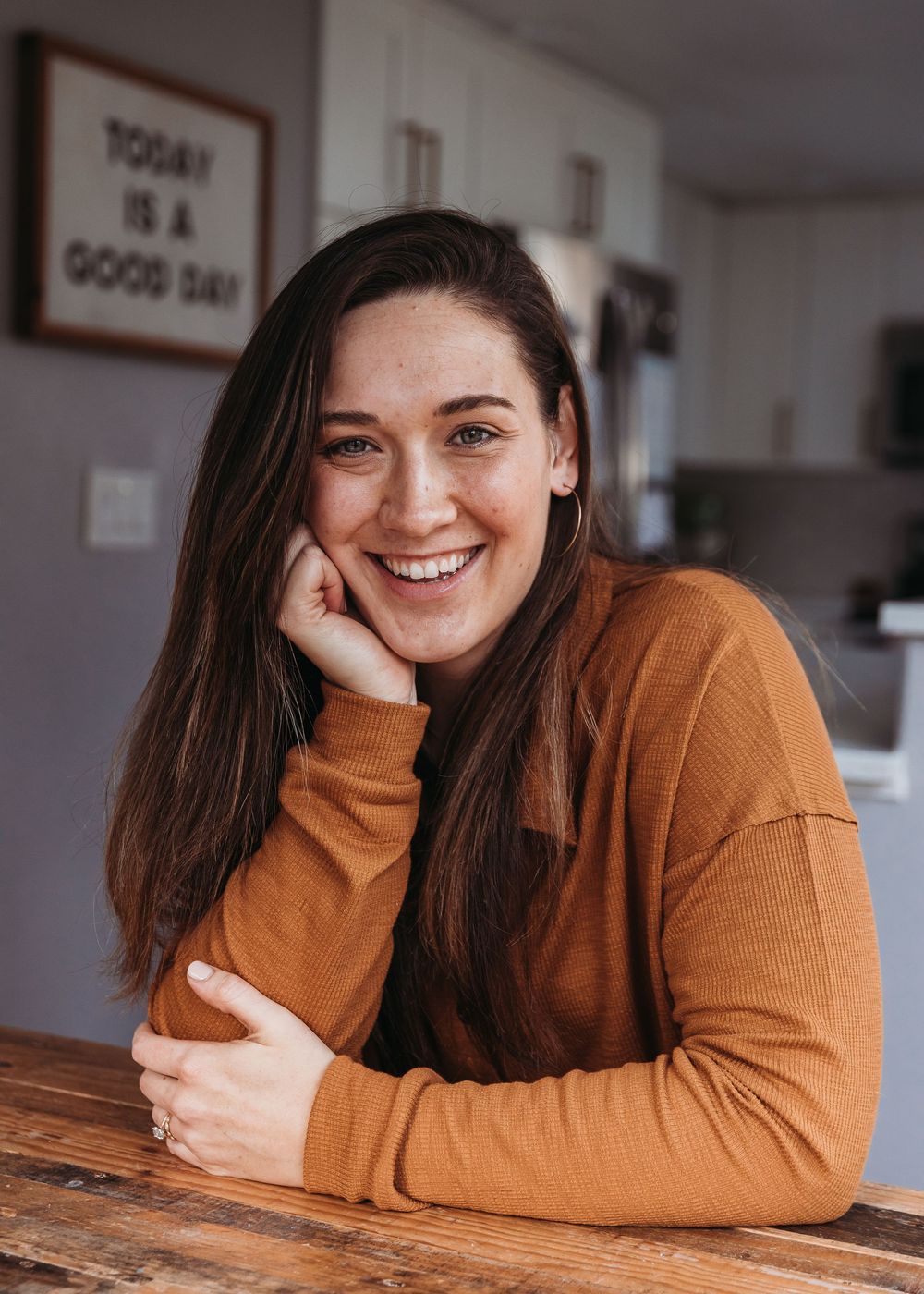 A person in a brown turtleneck sweater rests their head on their hand while smiling at a wooden table indoors.