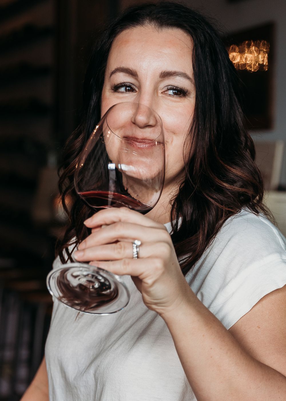 Someone holds a glass of red wine while smiling warmly in a casual setting with warm lighting in the background.