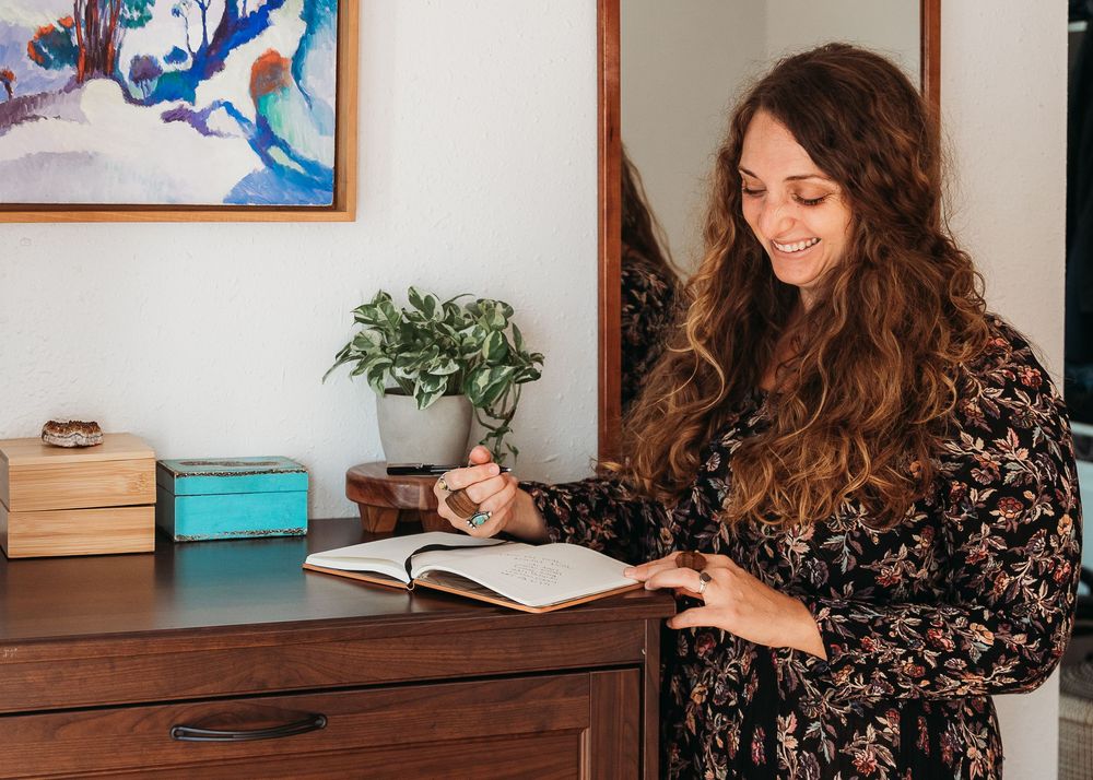 A person in a floral dress sits at a wooden desk with a journal, plant, and colorful wall art in the background.